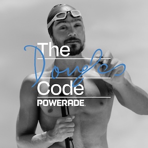 Zwart-witafbeelding van Douglas Matera met ontbloot bovenlichaam, zwemcap en zwembril, die een iets vasthoudt en met een marker iets in blauwe inkt schrijft over de tekst “The Code POWERADE”.