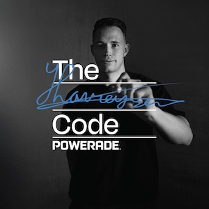 Zwart-witfoto van Harrie Lavreysen in een donker T-shirt die met een blauwe stift op het camerabeeld schrijft. Op de voorgrond staat de tekst “The Code POWERADE”.