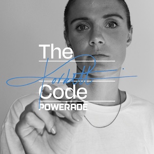 Zwart-witafbeelding van Kaylene Corbett in een T-shirt en ketting die met een marker iets in blauwe inkt schrijft over de tekst “The Code POWERADE”;.