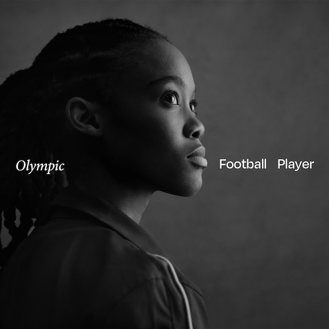Zwart-witfoto van Linda Motlhalo met lange, in vlechten gedragen haren, gekleed in een sportjack met strepen op de mouw. Op de afbeelding staat de tekst “Olympic Football Player” tegen een effen, donkere achtergrond.