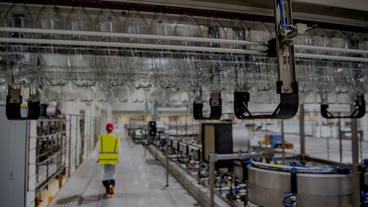 Een foto van de fabriek in Dongen waar je lege plastic flessen ondersteboven aan een transportrails ziet hangen. Op de achtergrond loopt een medewerker met een veiligheidsvest door de fabriekshal.